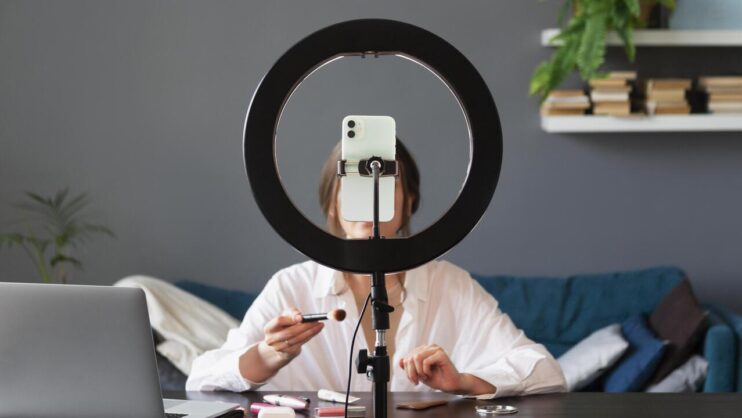 Influencerka nagrywająca kontent makijażowi, na pierwszym planie widoczna lampa pierścieniowa i telefon